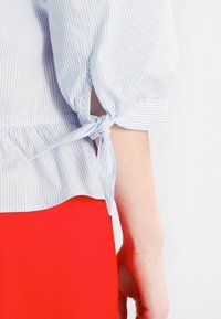 Chemise rayée bleu clair et blanc avec un détail noué aux manches et une taille froncée, complétée par une jupe unie rouge.