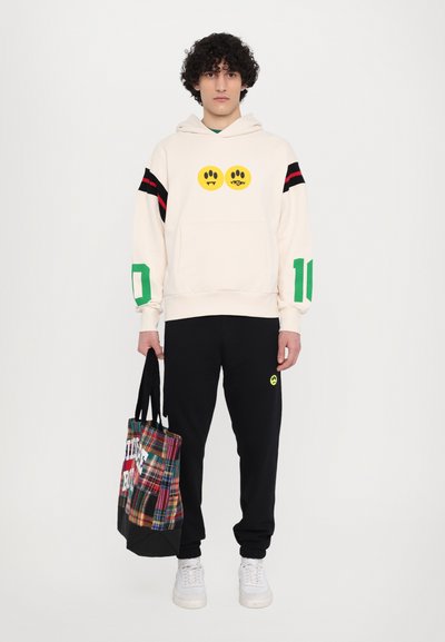 Beige hoodie with yellow smiley face graphic, red and black stripes on sleeves, green accents, paired with black sweatpants and plaid tote bag.