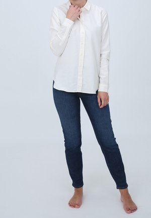 Personne debout pieds nus, portant une chemise blanche à boutons et un jean skinny bleu foncé, sur un fond clair uni.