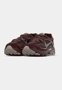 Zapatillas deportivas granate con parte superior de malla, detalles grises, suela gruesa de goma y puntera redondeada. Los cordones son marrones y la lengüeta lleva una marca.