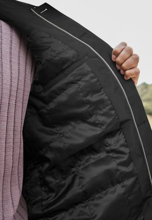 Veste noire isolée avec un intérieur matelassé, fermeture éclair argentée et une poche extérieure. Le tissu est lisse avec un fini mat.