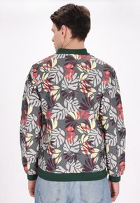 Bloemen bomberjack met een grijze basis en meerkleurige bladerenpatronen in rode, gele en groene accenten. Ribbelkraag en zoom in het groen.