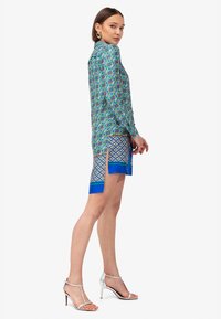 Blusa a fantasia nei toni del verde, blu e rosso con maniche lunghe, abbinata a pantaloni corti con un design geometrico e rifiniture blu, completata da décolleté argentati.