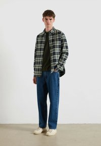 Camicia a quadratini in verde scuro, blu navy e beige, indossata sopra una maglietta verde; abbinata a jeans blu larghi e scarpe da ginnastica color crema.