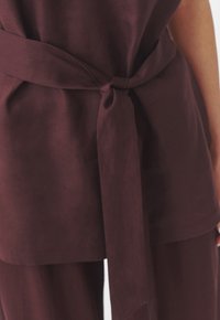 Maroon laneni top s vezanom strukom. Glatka tekstura, dizajn s kratkim rukavima, s čvorastim detaljem na struku i ravnim krojem.