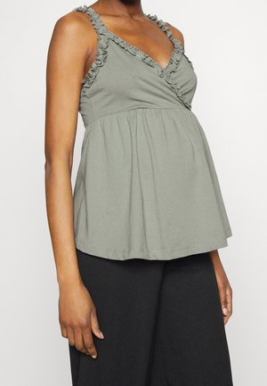 Femme portant un haut de maternité gris sans manches avec volants et un pantalon noir, montrée des épaules aux hanches.