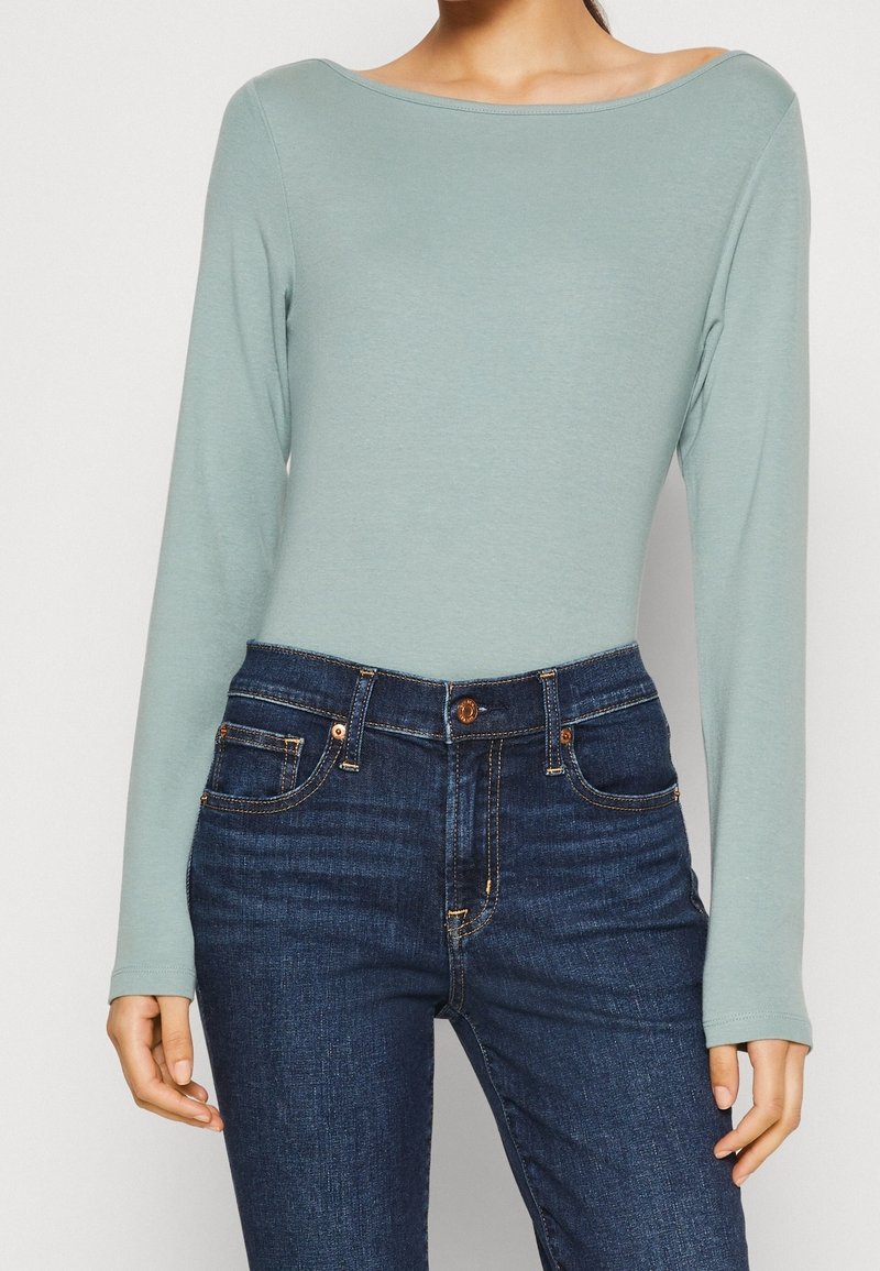 Jasnoniebieski top z długim rękawem i dekoltem w łódkę, w połączeniu z ciemnoniebieskimi dżinsami mającymi miedziane akcenty i pięć kieszeni.
