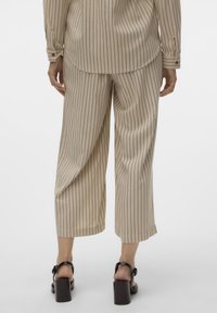 Persona che indossa pantaloni beige a gamba larga con righe sottili e una camicia a maniche lunghe abbinata, con sandali neri con tacco, vista da dietro.