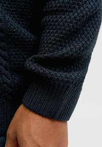 Knitted sweater in dark navy with a textured pattern. Features ribbed cuffs and a cable-knit design along the seam. Soft, thick fabric.