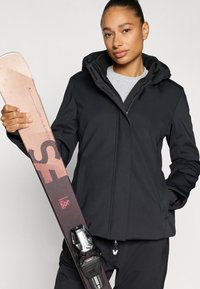 Čierna lyžiarska bunda s kapucňou, textúrovaná látka a zips na zapínanie, drží ružové a hnedé lyže s viazaním, na neutrálnom pozadí.