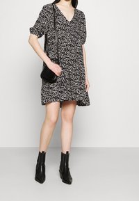 Mini-robe à motifs noirs et blancs avec un col en V, des manches bouffantes et une taille froncée. Associée à des bottines noires et un sac bandoulière texturé.