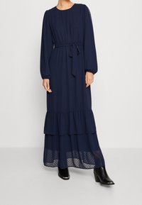 Granatowa sukienka maxi z długimi, bufkowatymi rękawami, talią podkreśloną paskiem oraz falbanami na stopniowanym dole. Posiada subtelną kropkowaną fakturę i przezroczystą dolną warstwę.