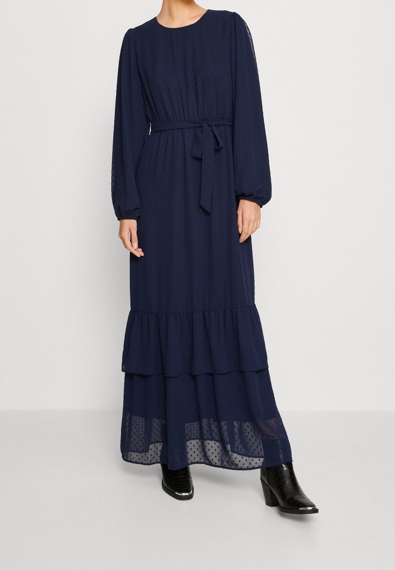 Granatowa sukienka maxi z długimi, bufkowatymi rękawami, talią podkreśloną paskiem oraz falbanami na stopniowanym dole. Posiada subtelną kropkowaną fakturę i przezroczystą dolną warstwę.