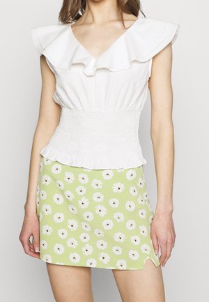 Vrouw draagt een wit mouwloos topje met ruches en een lichtgroene minirok met witte bloemen en een kleine zijsplit.