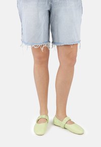 Ballerines en cuir vert clair avec un détail froncé à la cheville, portées avec des shorts en denim bleu clair effilochés, mettant en avant un style décontracté et relax.