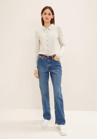 Top largo de manga larga en color beige claro con canalé, cuello y botones, combinado con jeans de pierna recta azules y zapatillas blancas, sobre un fondo neutro.