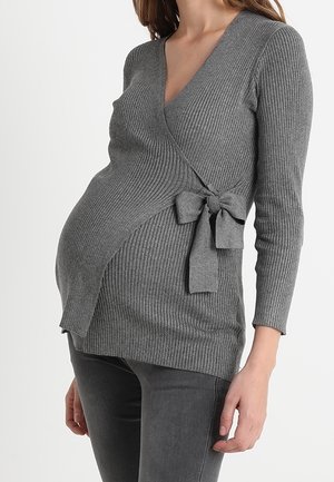 Zwangere persoon draagt een grijze geribbelde omslagtrui met een strik aan de zijkant, gecombineerd met donkergrijze broek tegen een effen achtergrond.