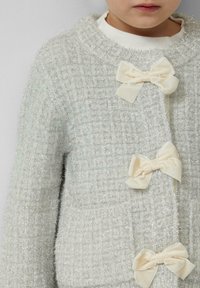 Jasnoszary kardigan o teksturowanej powierzchni z puszystym wykończeniem, z trzema kremowymi satynowymi kokardkami i okrągłym dekoltem.