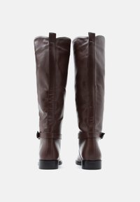 Καφέ δερμάτινες μπότες μέχρι το γόνατο με λεία υφή, διακοσμημένες με λουρί, επίπεδη φτέρνα και ορατή ραφή κατά μήκος της πίσω ραφής.