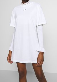 White Nike t-shirt dress with short sleeves and a mock neckline. Features a small black logo on the chest and ruffled cuffs on the inner sleeves.