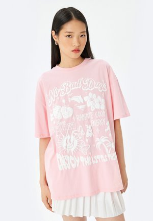 Camiseta rosa de gran tamaño con impresión gráfica blanca que presenta texto e ilustraciones de flores, mariposas y motivos de sol. Mangas cortas, cuello redondo.