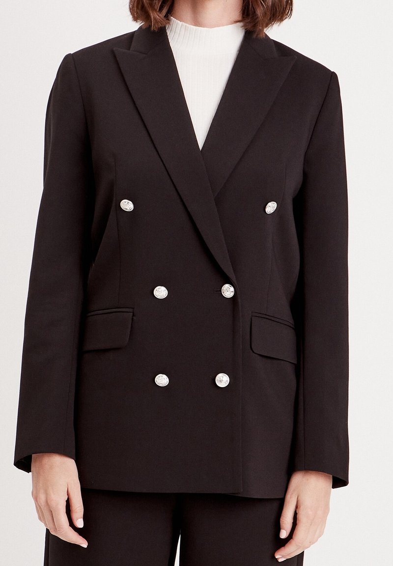 Femme portant un blazer noir à double boutonnage avec des boutons argentés, sur un top blanc côtelé à col montant, sur un fond uni.