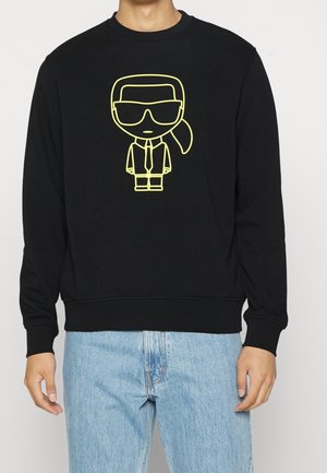Mand iført sort sweatshirt med gul tegneseriefigur iført solbriller og slips, kombineret med lyseblå jeans.