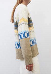 Femme portant un pull en tricot à motifs crème et beige avec des designs géométriques bleus, jaunes et marron, associé à un pantalon blanc.
