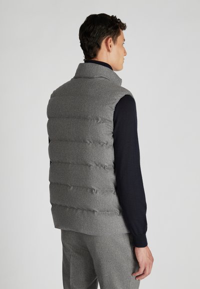 Mann in grauem, gestepptem ärmellosen Weste über einem schwarzen Langarmshirt und grauen Hosen, blickt von hinten vor einem schlichten Hintergrund.