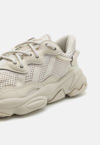 Baskets beige clair avec une tige en mesh, des panneaux texturés et des lacets ton sur ton. La semelle présente un design ondulé avec des détails de traction bien visibles.