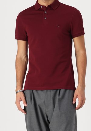 Mann trägt ein bordeauxrotes Polohemd mit kurzen Ärmeln und Knöpfen sowie eine graue Hose und steht vor einem schlichten, hellen Hintergrund.