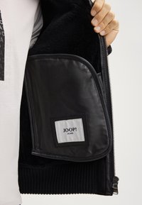 Giacca nera con zip e fodera morbida e testurizzata. Presenta una tasca in pelle con etichetta del logo. Il polsino a coste all'orlo aggiunge un tocco di dettaglio.