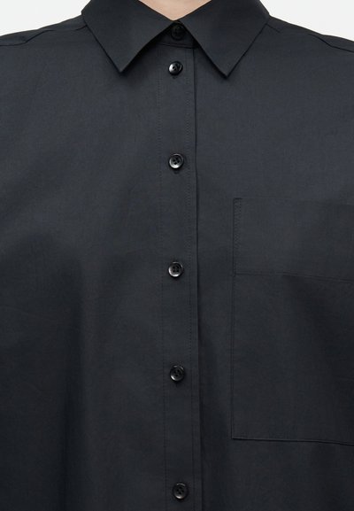 Μαύρο πουκάμισο με κουμπιά, με αιχμηρό γιακά και τσέπη στο στήθος, εμφανιζόμενο στο άνω μέρος του κορμού ενός ατόμου.