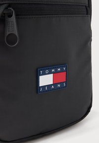 Lähikuva mustasta kangaskassista, jossa on etupuolella vetoketju ja suorakaiteen muotoinen logopläkki, jossa lukee "Tommy Jeans" valkoisella, punaisella ja sinisellä värillä.