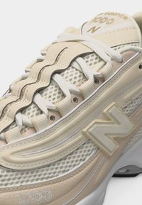 Gros plan sur une basket New Balance 1000 beige et blanche avec des panneaux en mesh, un revêtement en cuir ondulé distinctif, et des lacets blancs.