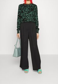 Pull imprimé léopard vert, pantalon large à carreaux noirs, sac à main bleu sarcelle, et sandales à bride nouée bleu sarcelle. Textures lisses, motifs audacieux.