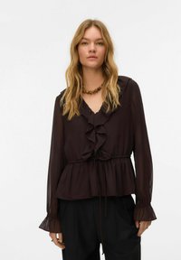 Femme aux longs cheveux blonds portant un chemisier marron à volants avec des manches longues transparentes et un collier de perles, debout devant un fond uni.