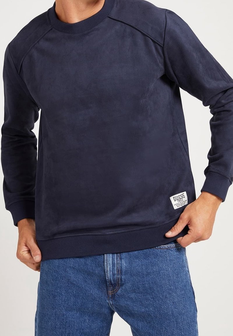 Homme portant un sweat-shirt bleu marine à manches longues avec un petit patch du logo Guess blanc sur le bas à gauche, associé à un jean bleu.