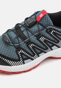 Scarpa da atletica leggera con tomaia in mesh blu, dettagli neri, strisce riflettenti e un linguetta rossa, dotata di un sistema di allacciatura sicuro.