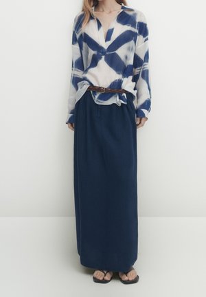 Woman wearing a loose blue and white tie-dye shirt tucked into a navy long skirt with a brown braided belt and black sandals.