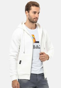 Witte rits-hoodie met een voorkant zak en een trekkoordkap, gelaagd over een wit T-shirt met een kleurrafische afbeelding en zwarte tekst.