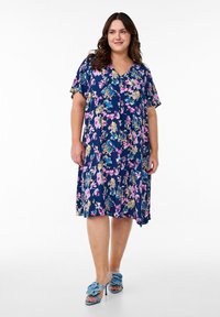 Robe florale marine à manches courtes et décolleté en V. Présente des motifs roses, bleus et dorés. Portée avec des sandales à talons bleu clair.