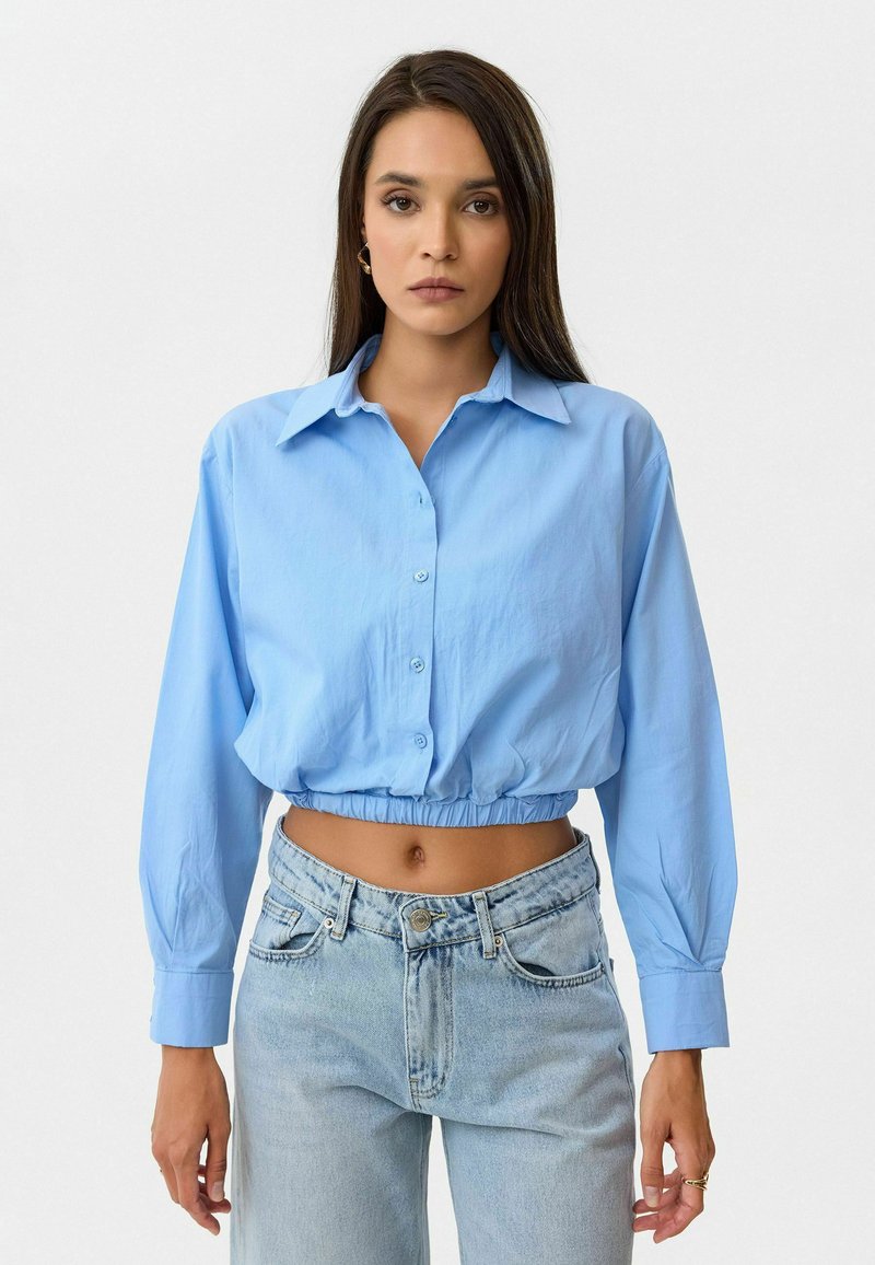 Lichtblauwe cropped shirt met een knoopsluiting aan de voorkant, lange mouwen en elastische zoom, gecombineerd met hoge taille vervaagde blauwe jeans.