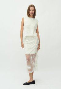 Top sin mangas de texturizado blanco combinado con una falda blanca que presenta bordado floral en una sobrecapa transparente. La modelo lleva flats negros.