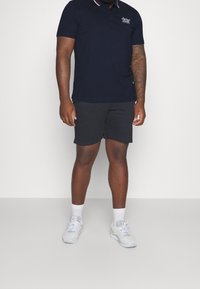 Marinblå piké med röd och vit krage, logotyp på bröstet, matchad med mörka shorts och vita sneakers med grå detaljer.