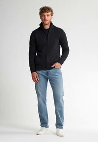 Schwarzer Reißverschluss-Pullover mit geripptem Muster und hohem Kragen, kombiniert mit hellblauen Jeans und weißen Turnschuhen. Modell steht frontal zur Kamera.