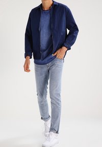 Granatowa kurtka zapinana na zamek over blue shirt, zestawiona z jasnoniebieskimi dżinsami i białymi trampkami. Kurtka ma luźny krój i ryflowane mankiety.