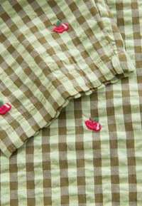 Roheline ja pruun ruuduline kangas tikitud punaste õuntega, millel on pehme tekstuur ja kerge materjal. Muster on mänguline ja dekoratiivne.