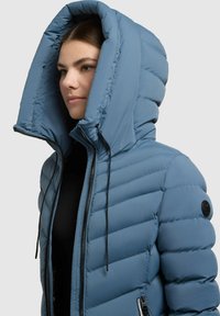 Sieviete ar zilu polsterētu jaku ar lielu kapuci un melnu rāvējslēdzēju, vērsta pa kreisi uz vienkārša fona.