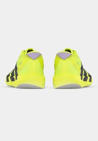 Αθλητικά παπούτσια νέον κίτρινου με μαύρες ρίγες, λαστιχένιες σόλες και λοξά λιλά στοιχεία στη φτέρνα. Η υφή φαίνεται λεία και ελαφριά.